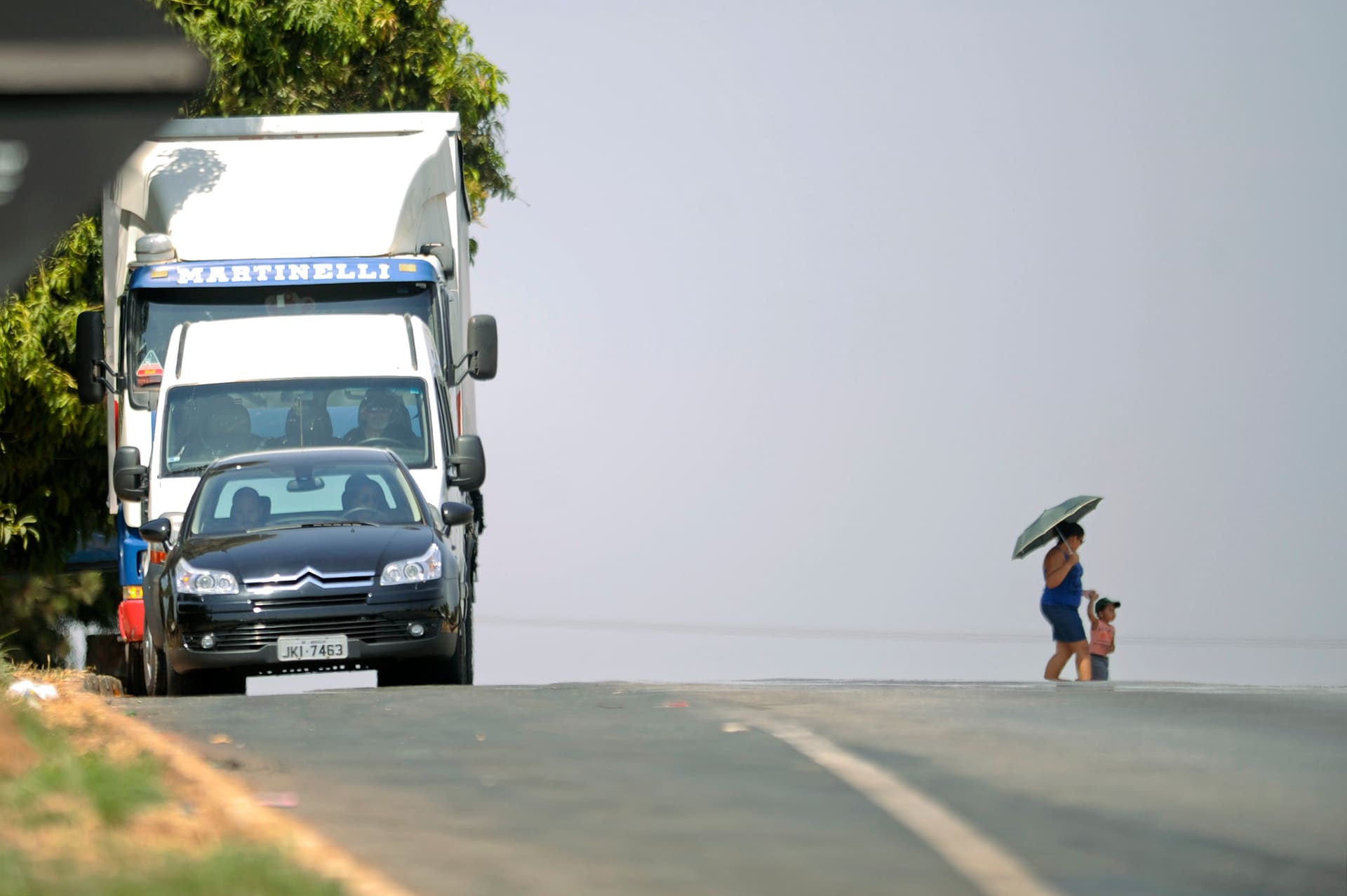 Caminhão e carro trafegam por rodovia federal enquanto mulher e criança caminham pelo acostamento